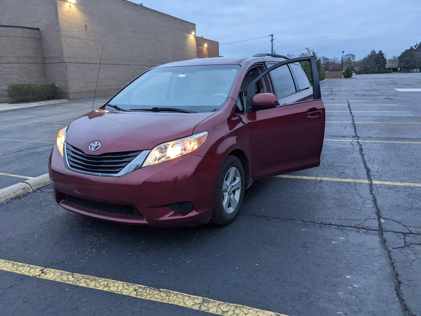 A dark red 2014 Toyota Sienna minivan parked in an empty lot at dusk with its headlights on and a sliding door open.